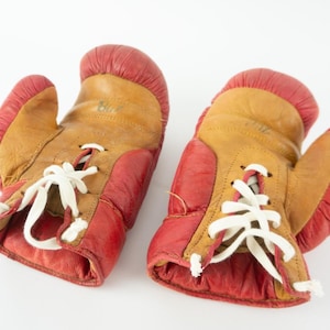 May include: A pair of vintage boxing gloves. The gloves are primarily tan with red accents on the cuffs and thumb. White laces secure the gloves. The gloves are likely made of leather.