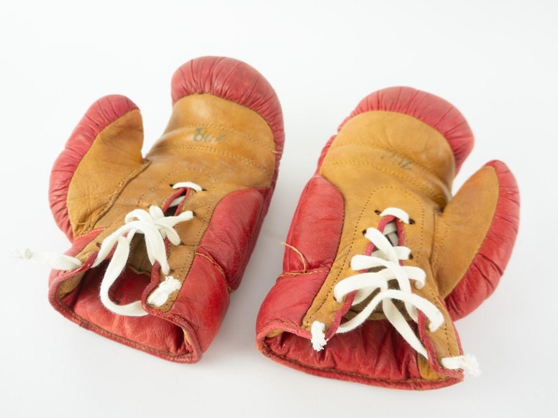 May include: A pair of vintage boxing gloves. The gloves are primarily tan with red accents on the cuffs and thumb. White laces secure the gloves. The gloves are likely made of leather.