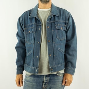 Peut inclure: Veste en jean bleue avec des boutons, des poches de poitrine et un col. La veste est portée sur un t-shirt gris et associée à un jean bleu. Le style général est décontracté et classique.