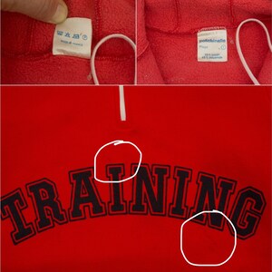 K&ouml;nnte beinhalten: Roter Fleece-Pullover mit einem schwarzen "TRAINING"-Aufdruck auf der Vorderseite. Der Pullover hat einen wei&szlig;en Rei&szlig;verschluss und ein Etikett mit der Aufschrift "polticinelle" mit dem Text "68% Baumwolle 32% Polyamid".