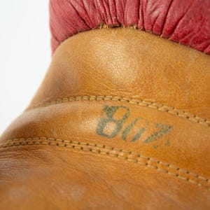 May include: Close-up of a vintage boxing glove. The glove is made of tan leather with a red leather cuff. The number "807" is stenciled in green ink on the leather. The stitching is visible.