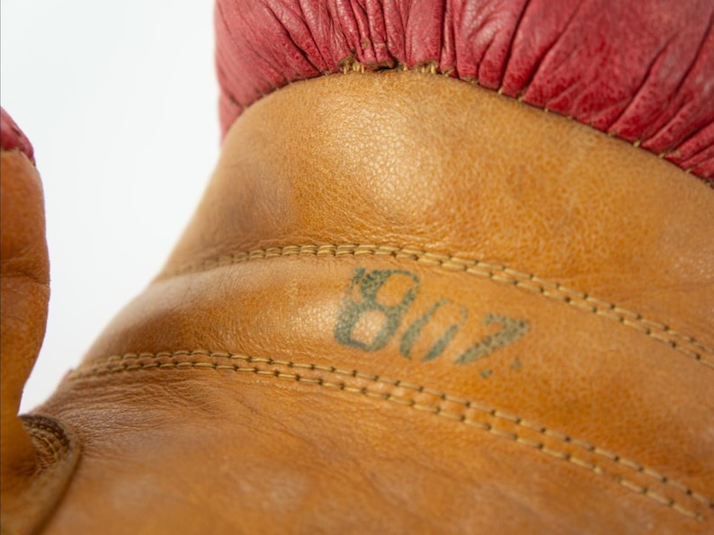 May include: Close-up of a vintage boxing glove. The glove is made of tan leather with a red leather cuff. The number "807" is stenciled in green ink on the leather. The stitching is visible.