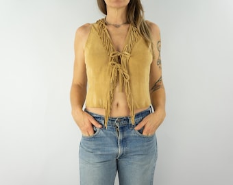 May include: A brown suede vest with fringe detailing. The vest has a tie closure at the front and is sleeveless. The model is wearing blue jeans.