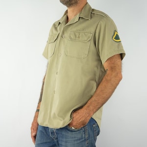 Peut inclure: Une chemise boutonnée à manches courtes de couleur kaki avec un écusson sur la manche gauche. L'écusson est vert et jaune avec un motif géométrique.