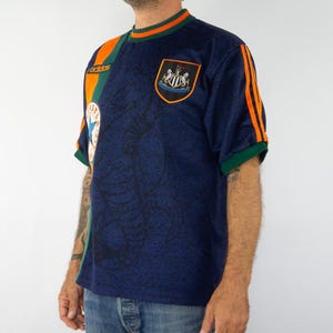 May include: Navy blue soccer jersey with orange and green accents. The jersey features the Adidas logo, a team crest, and orange stripes on the sleeves. The shirt has a patterned design.