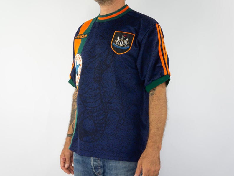 May include: Navy blue soccer jersey with orange and green accents. The jersey features the Adidas logo, a team crest, and orange stripes on the sleeves. The shirt has a patterned design.