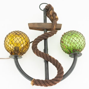 May include: A wall-mounted anchor-shaped lamp with two glass globes. The globes are covered in a net-like material and are yellow and green. The anchor is made of metal and has a rope wrapped around it.