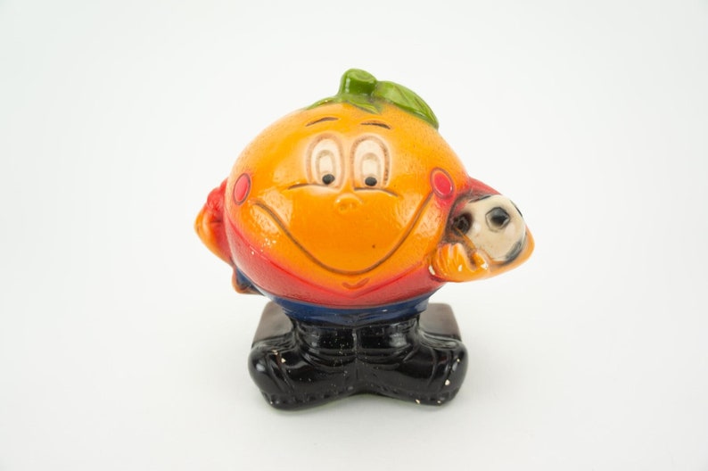 May include: A ceramic figurine of an orange with a smiling face wearing a red and blue soccer uniform and holding a soccer ball.