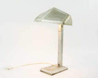 May include: A silver-toned metal desk lamp featuring a rectangular base, a vertical support arm, and a wide, angled shade with textured panels. The lamp includes a thin, light-colored power cord extending from the base with an inline switch.