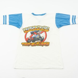 以下が含まれることがあります： 白のTシャツ、青い袖とクルーネック。赤い円の中にレーシングカーのグラフィックがあり、「NITROTERS」と「THE OUTLAWS」の文字が黄色とオレンジ色で描かれています。