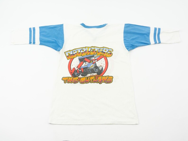 以下が含まれることがあります： 白のTシャツ、青い袖とクルーネック。赤い円の中にレーシングカーのグラフィックがあり、「NITROTERS」と「THE OUTLAWS」の文字が黄色とオレンジ色で描かれています。