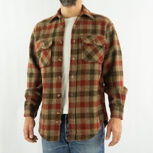 Puede incluir: Una camisa abotonada de manga larga con un estampado a cuadros rojo, verde y marrón. La camisa tiene dos bolsillos en el pecho y un cuello. El modelo lleva una camiseta blanca debajo y vaqueros azules.