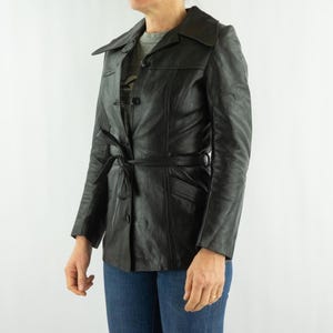 May include: Black leather jacket with a belted waist and button closure. The jacket has a classic collar and long sleeves. The jacket is worn with blue jeans.