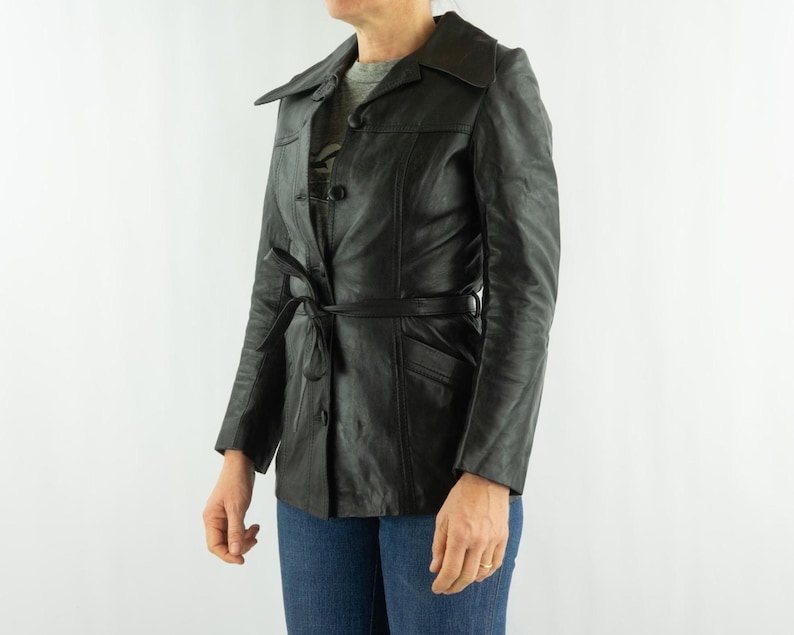 May include: Black leather jacket with a belted waist and button closure. The jacket has a classic collar and long sleeves. The jacket is worn with blue jeans.