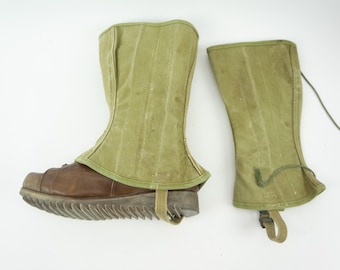 Könnte beinhalten: Ein Paar olivgrüne Canvas-Gamaschen mit vertikalen Nähten, wobei eine an einem braunen Lederschuh mit dicker, geriffelter Sohle und beigem Riemen befestigt ist, während die andere flach mit einer dünnen grünen Kordel und einem kleinen Schnallenriemen liegt.