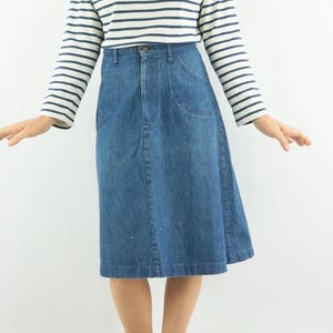 May include: A blue denim skirt with a classic design. The skirt has a button and zipper closure, front pockets, and a knee-length hemline. The model is wearing a striped long-sleeve shirt.