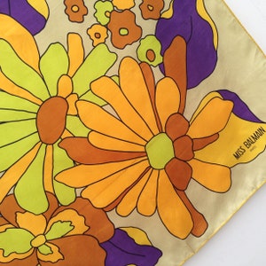 May include: A square silk scarf featuring a bold floral design in orange, brown, green, and purple on a cream backdrop. The scarf is labelled "MISS BALMAIN PARIS" in one corner.