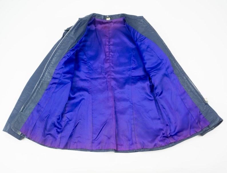 K&ouml;nnte beinhalten: Eine dunkelblaue Lederjacke mit einem leuchtend lila Satinfutter. Die Jacke ist offen und zeigt den Kontrast zwischen Au&szlig;en- und Innenseite. Das Futter hat einen Farbverlauf, der von Lila zu einem tieferen Farbton &uuml;bergeht.