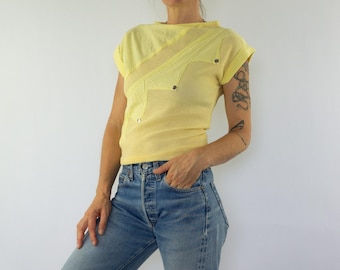Puede incluir: Una camiseta de manga corta en color amarillo pálido con una base de tejido de malla texturizada y paneles de tela lisa dispuestos en un patrón diagonal superpuesto. La prenda incluye pequeños remaches metálicos plateados en las costuras.