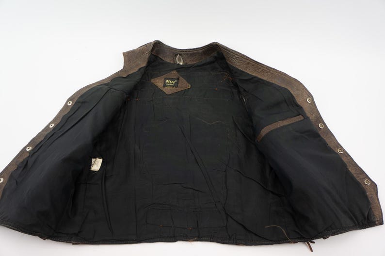 May include: Open view of a brown leather vest with a black interior lining. The vest has a button-up front, two interior pockets, and a small tag. The leather has a distressed appearance.