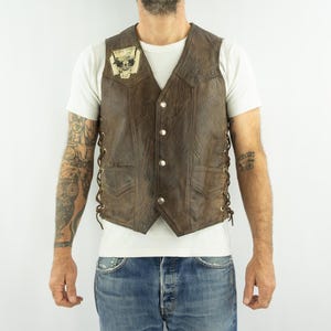 May include: Brown leather vest with a V-neck, button closure, and side lacing. Features a small patch on the chest. The vest has two front pockets and a textured finish. A classic biker style.