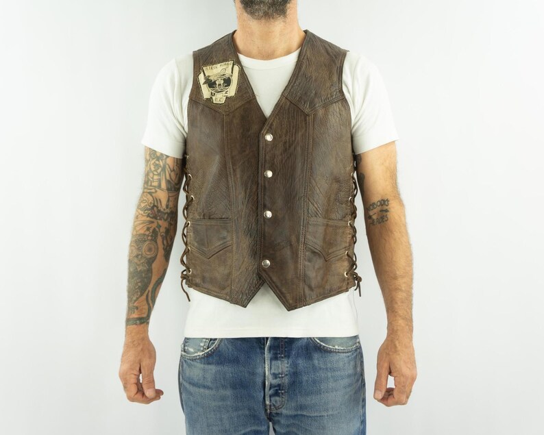 May include: Brown leather vest with a V-neck, button closure, and side lacing. Features a small patch on the chest. The vest has two front pockets and a textured finish. A classic biker style.