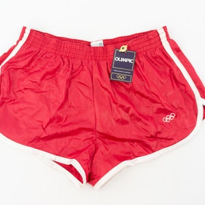 May include: Red athletic shorts with white trim and the Olympic rings logo. The shorts have an elastic waistband and a tag that reads "Olympic".