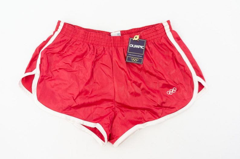 May include: Red athletic shorts with white trim and the Olympic rings logo. The shorts have an elastic waistband and a tag that reads "Olympic".