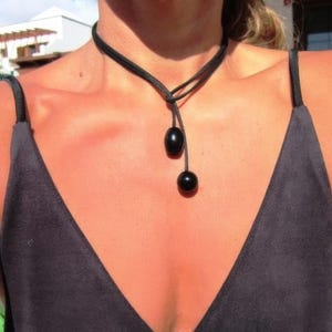 May include: A black cord necklace featuring two smooth, round black beads suspended from a simple knot. The cord wraps around the neck and hangs down the center of the chest, with the beads positioned at slightly different lengths for a layered look.