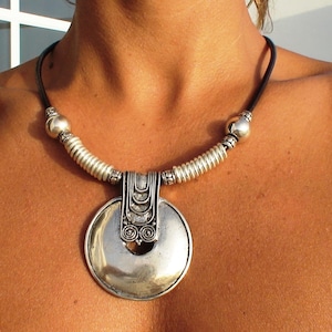 May include: A necklace featuring a black cord and a large, circular silver-toned pendant. The pendant is attached to the cord by a textured, rectangular silver-toned bail. The cord is accented with silver-toned beads and ribbed cylindrical spacers.