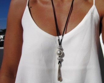 May include: A long necklace featuring a black cord, a large silver-toned spherical bead, two smaller textured silver-toned beads, and a smooth, elongated silver-toned pendant. The necklace is worn over a white sleeveless top with thin straps.