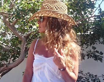 May include: A straw hat with a wide brim and open weave design. The hat is tan and worn by a person wearing sunglasses and a white top. The background includes a tree with green leaves and a white wall.