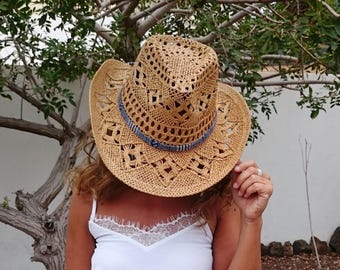 Puede incluir: Sombrero de vaquero de paja tejida con una banda azul decorativa. El sombrero tiene un diseño de tejido abierto y un ala ancha. El sombrero es de color marrón claro y es un accesorio de verano.