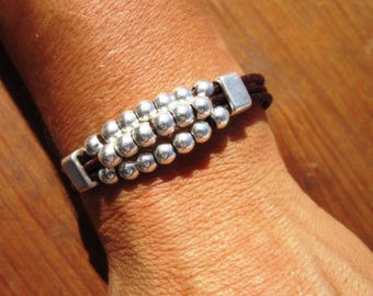 May include: A bracelet featuring three dark brown leather cords held together by two rectangular silver-toned metal end caps. Between the end caps, the leather cords are adorned with three parallel rows of small, round, polished silver-toned beads.