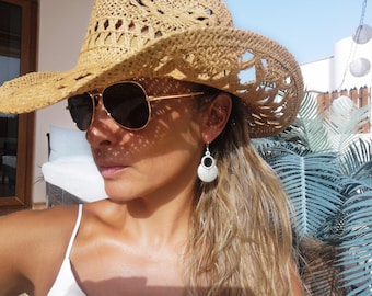 May include: A tan woven straw hat with a wide brim and cutout details, paired with dark-tinted aviator sunglasses featuring thin gold-tone frames and silver-tone drop earrings consisting of a small ring attached to a larger circular disc.