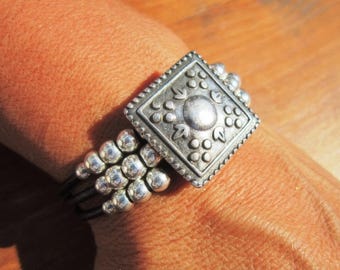 May include: A silver-toned ring featuring a square centerpiece with an embossed floral and dot pattern. The band consists of three thin dark wires adorned with multiple small, round, polished silver-toned beads arranged in a cluster on the side.