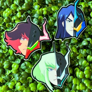 Könnte beinhalten: Drei bunte Aufkleber mit Anime-Charakteren, deren Gesichter zur Hälfte von einem schwarzen und blauen mechanischen Design verdeckt werden. Die Charaktere sind eine Frau mit grünen Haaren, eine Frau mit blauen Haaren und ein Mann mit roten und schwarzen Haaren.