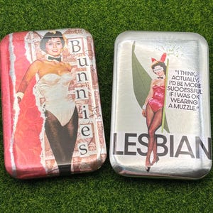 May include: Two rectangular metal tins with vintage-style images. One tin features a woman in a bunny costume with the word "Bunnies". The other tin has a woman in a pink bunny costume with the word "Lesbian" and a quote.