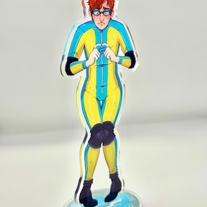 May include: An acrylic standee featuring a cartoon character with red hair and glasses, wearing a yellow and blue full-body suit with black knee pads and black boots. The character has a worried expression and is standing on a blue puddle base.