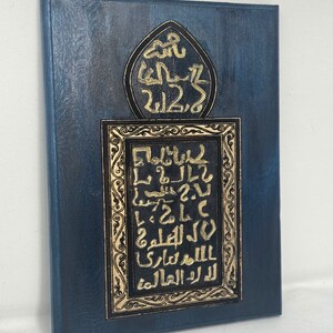 May include: A blue wooden plaque with gold Arabic calligraphy. The calligraphy is framed in a gold ornate border. The plaque is a decorative piece of art.