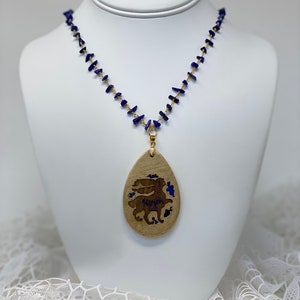 May include: A gold necklace with a blue stone pendant. The pendant is teardrop-shaped and features a gold-coloured design of a rabbit inside. The necklace is made of small, irregular blue stones.