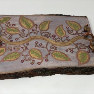 May include: A wooden serving tray with a natural bark edge, painted with a whimsical design of a vine with leaves and flowers in shades of green, gold, and purple. The tray has two copper leaf-shaped handles.