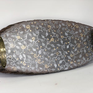 May include: A wooden serving tray with a grey and gold leaf design. The tray has two brass handles on the sides.