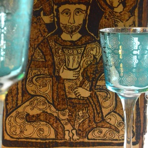 May include: A woodburned image of a king sitting on a throne, holding a cup. The image is framed in a wooden frame. Two teal blue glasses are in the foreground.