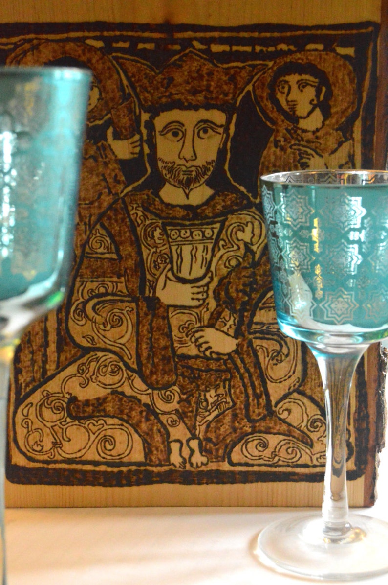 May include: A woodburned image of a king sitting on a throne, holding a cup. The image is framed in a wooden frame. Two teal blue glasses are in the foreground.