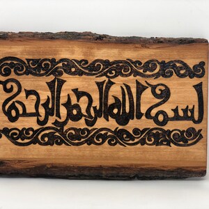 Puede incluir: Un letrero de madera con un borde de corteza natural, que presenta un diseño quemado en negro de caligrafía árabe con adornos decorativos. El texto dice "سبحان الله والحمدلله"