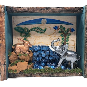 May include: A small wooden box with a blue painted frame, containing a miniature scene with a gold rabbit and a silver elephant facing each other. The scene is set against a backdrop of blue and green painted foliage and a blue crescent moon.