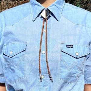 May include: A light blue button-down shirt with a brown leather bolo tie. The shirt has a Wrangler logo on the pocket.
