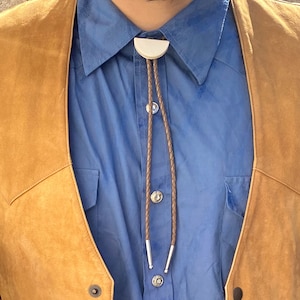May include: A person wearing a blue button-down shirt with a brown leather bolo tie. The shirt has a white collar and the bolo tie has a silver clasp. The person is also wearing a brown suede vest.