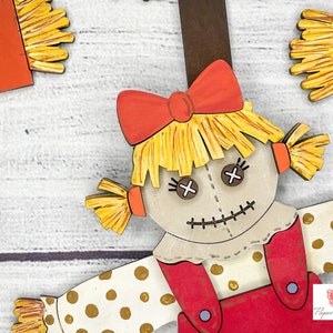 May include: A wooden cutout of a scarecrow with a red and white polka dot dress, a red bow, and a straw hat. The scarecrow has a stitched smile and button eyes.